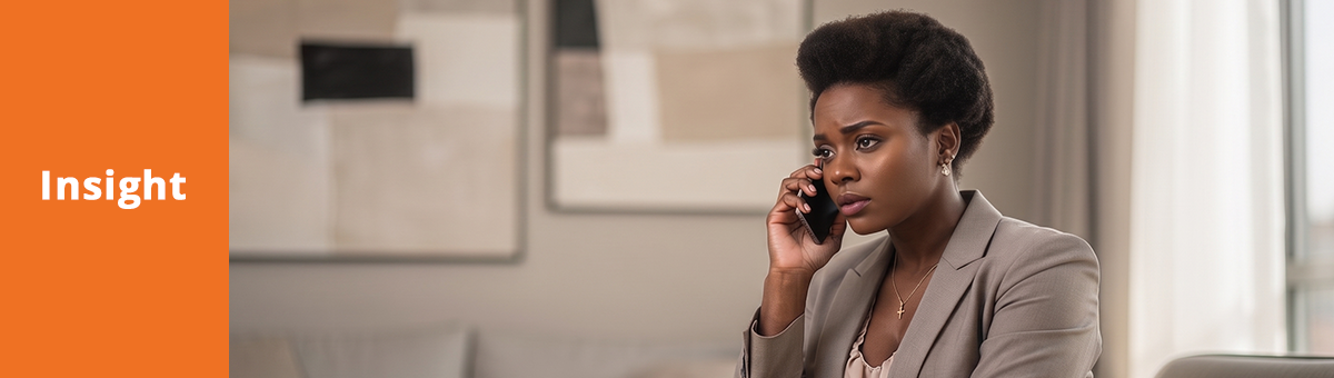 A woman sits in a living room, holding a smartphone to her ear. She has an annoyed facial expression.